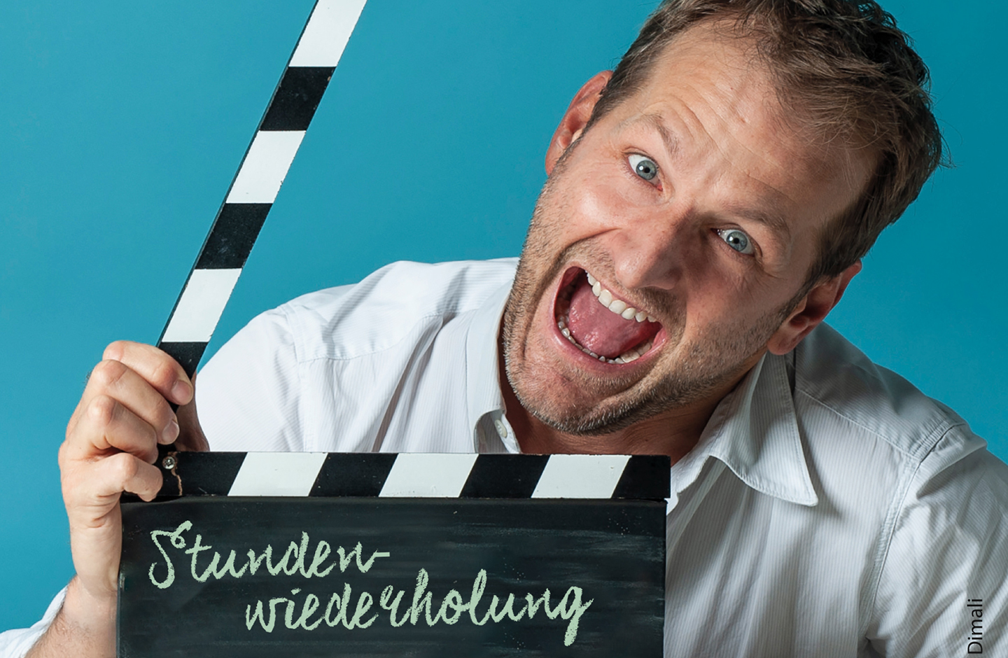 Der Kabarettist Andreas Ferner hält eine Film-Klappe in der Hand auf der "Stundenwiederholung" steht